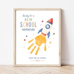 Könnte beinhalten: Ein gerahmter Druck mit einer Aquarell-Rakete und einer gelben Handabdruck-Abgasfahne. Der Text lautet Ready for a NEW SCHOOL ADVENTURE und FIRST DAY OF SCHOOL 2026. Kleine bunte Sterne sind um die Rakete herum auf weißem Grund verteilt.