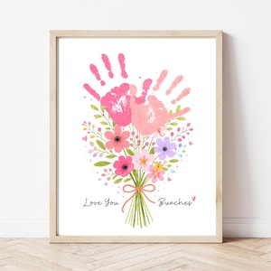 Può includere: Stampa incorniciata con un bouquet di fiori e due impronte di mani rosa. Le parole "Love You Bunches" sono scritte sotto. L'opera d'arte è su sfondo bianco, con una cornice in legno.