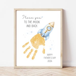 May include: A framed print with a light wood frame. The print features a blue rocket ship and a yellow handprint, with the text "I love you to the moon and back." Below is "Happy Father's Day 2026."