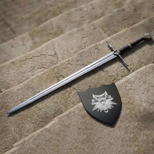 May include: A silver sword with a black handle and crossguard lies on stone steps beside a black shield. The shield displays a white wolf emblem. The sword has a long, straight blade.