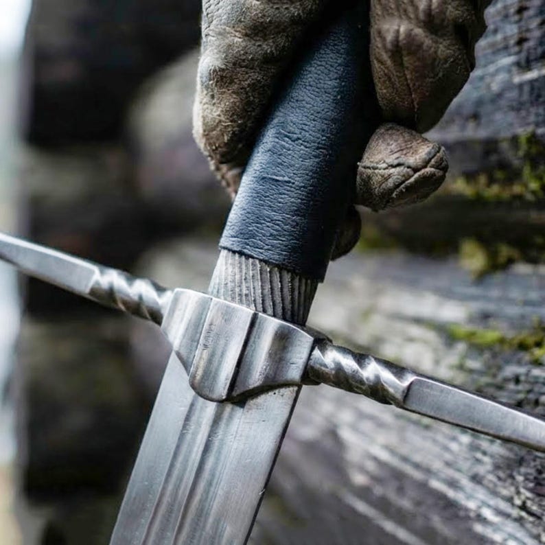 May include: A close-up shot of a medieval sword. The silver blade contrasts with the black leather-wrapped handle. The hilt features a twisted design. The sword is held by a gloved hand, evoking a historical or fantasy setting.