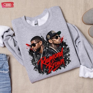 Digital bootleg rap tee graphic featuring R&B artists Usher and Chris Brown standing back-to-back wearing sunglasses and black jackets. The background showcases a high-contrast nighttime city skyline with glowing lights, abstract geometric stars, and bold red brushstroke text reading "Raymond & Brown".