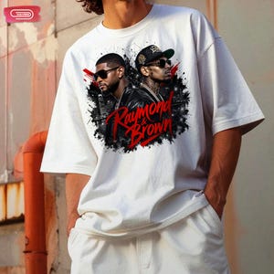 Digital bootleg rap tee graphic featuring R&B artists Usher and Chris Brown standing back-to-back wearing sunglasses and black jackets. The background showcases a high-contrast nighttime city skyline with glowing lights, abstract geometric stars, and bold red brushstroke text reading "Raymond & Brown".