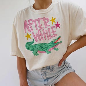 Detailed teal and pink crocodile illustration with "AFTER WHILE" blue hand-lettered graffiti font and green stars, trending boy boutique design for kids' apparel.