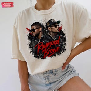 Digital bootleg rap tee graphic featuring R&B artists Usher and Chris Brown standing back-to-back wearing sunglasses and black jackets. The background showcases a high-contrast nighttime city skyline with glowing lights, abstract geometric stars, and bold red brushstroke text reading "Raymond & Brown".