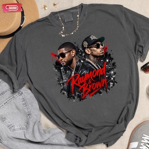 Digital bootleg rap tee graphic featuring R&B artists Usher and Chris Brown standing back-to-back wearing sunglasses and black jackets. The background showcases a high-contrast nighttime city skyline with glowing lights, abstract geometric stars, and bold red brushstroke text reading "Raymond & Brown".