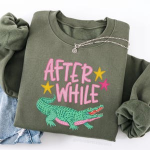 Detailed teal and pink crocodile illustration with "AFTER WHILE" blue hand-lettered graffiti font and green stars, trending boy boutique design for kids' apparel.