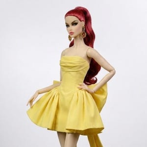 May include: A fashion doll wearing a strapless yellow mini dress with a large bow in the back and a long sash. The doll has red hair, gold earrings, and beige high-heeled booties. The doll is posed against a white background.