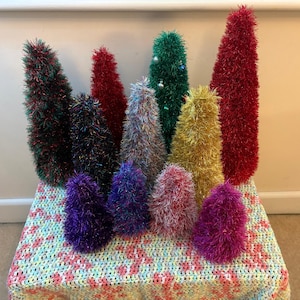 Puede incluir: Una colección de árboles de Navidad decorativos y coloridos de varios tamaños. Los árboles están hechos de un material difuso similar al oropel en tonos de rojo, verde, dorado, morado y plateado. Están colocados sobre una manta de ganchillo.