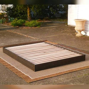 May include: A dark brown wooden bed frame with wooden slats. The bed frame is rectangular and sits on a brown and beige rug. The bed frame is outdoors on a concrete surface.