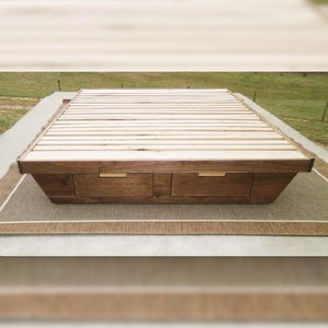 May include: A low-profile wooden bed frame with two drawers. The frame is made of dark brown wood with a light-colored wooden slat platform. The drawers have gold-colored handles. The bed frame is placed on a neutral-colored rug.