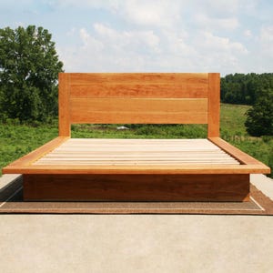 May include: A wooden bed frame with a headboard. The bed frame is made of light brown wood and has a simple, modern design. The headboard is rectangular and the bed frame sits on a low platform. The bed frame is empty, with wooden slats visible.