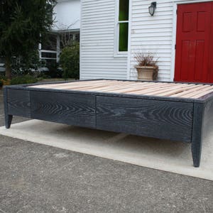 May include: A dark gray wooden bed frame with a slatted headboard and footboard. The frame has two drawers and sits on four legs.