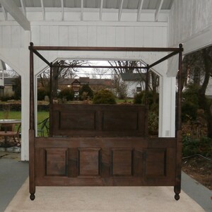 May include: A dark brown wooden canopy bed with four poster posts and a paneled headboard and footboard. The bed is in a white room with a view of trees and houses through a window.