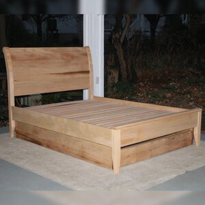 May include: A wooden sleigh bed frame with a built-in drawer. The bed frame is made of natural wood with a light brown finish. The drawer is located underneath the bed frame and is also made of wood.