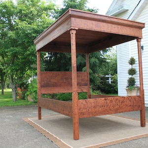 May include: A wooden canopy bed frame with a dark brown finish. The bed frame has a simple design with four posts and a flat headboard. The bed is sitting on a brown rug in front of a white building with a red door.