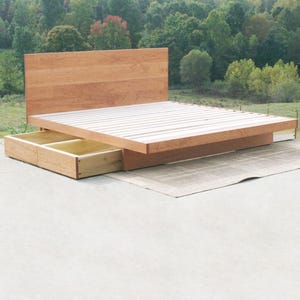 May include: A wooden platform bed with a headboard and two pull-out drawers. The bed frame and headboard are made of light brown wood. The bed sits on a concrete surface with a natural background of trees.