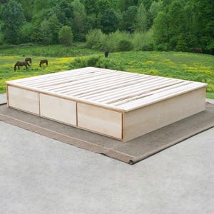 May include: A light-colored wooden platform bed with three drawers. The bed frame has a slatted top and is set on a neutral-colored rug. The background features a green field and trees.