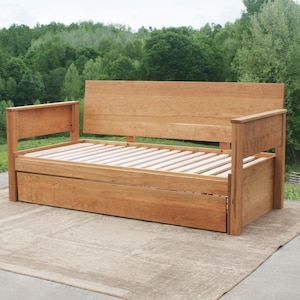 May include: A wooden daybed with a pull-out drawer. The daybed is made of solid wood and has a natural finish. The daybed is in a natural setting with trees in the background.
