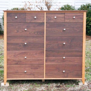 May include: A tall wooden dresser with a dark brown finish and light brown trim. The dresser has multiple drawers with silver knobs. The wood grain is visible on the drawer fronts. The dresser is standing on a grassy surface.