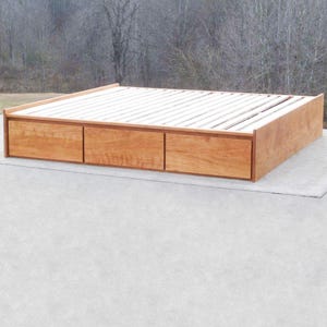 May include: A wooden platform bed frame with three drawers. The bed frame is made of light brown wood and has a slatted headboard.