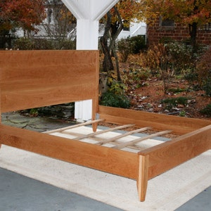 May include: A wooden bed frame with a slatted headboard and footboard. The frame is made of cherry wood and has a natural finish. The bed is sitting on a white rug.