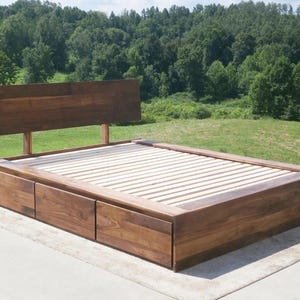 May include: A wooden bed frame with a headboard and two storage drawers. The bed frame is made of dark brown wood and has a slatted base. The headboard is rectangular and is attached to the frame with two wooden supports.