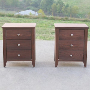 May include: Two matching dark brown wooden nightstands with three drawers each. The nightstands have silver knobs and are set against a blurred outdoor background. The tops of the nightstands are a lighter shade of brown.
