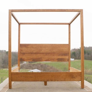 May include: A wooden canopy bed frame with a simple, rectangular design. The frame is made of natural wood with a light brown finish. The bed frame is standing on a light brown surface.