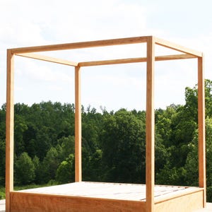 May include: A wooden canopy bed frame with a natural finish. The bed frame has a rectangular base and a four-post canopy structure. The bed is set outdoors with a backdrop of green trees and a blue sky.