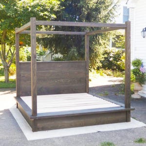 May include: A dark stained wooden canopy bed frame. The bed has a headboard and a low-profile footboard. The frame is constructed with vertical posts and horizontal beams, creating a canopy structure. The bed frame is sitting on a white sheet.