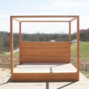 May include: A wooden canopy bed frame with a natural finish. The bed features a headboard and a slatted base. The frame is constructed with square posts and horizontal beams, creating a modern, minimalist design. The bed is outdoors on a concrete surface.