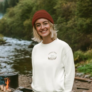 Könnte beinhalten: Ein cremefarbenes Sweatshirt mit dem Text "Green Trail Gear" und einer Berg-Grafik. Die Person im Bild trägt eine weinrote Mütze. Der Hintergrund zeigt einen Fluss und Bäume.