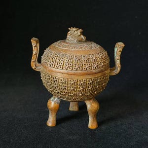 May include: An antique bronze incense burner with a lid, three legs, and two handles. The spherical body is intricately carved with a repeating pattern. The lid features a small dragon-like figure. The burner is set against a dark background.