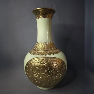 May include: A cream-colored vase with gold accents and a detailed dragon relief. The vase has a bulbous body, a slender neck, and a gold rim. The dragon design is prominently displayed on the front of the vase.