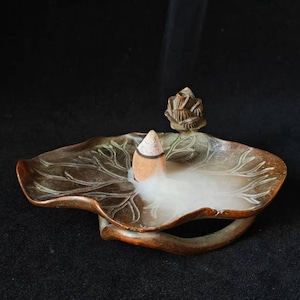May include: A brown, leaf-shaped incense holder with a carved floral accent. A lit incense cone releases white smoke, filling the holder. The holder has a detailed leaf vein design and a curved handle.