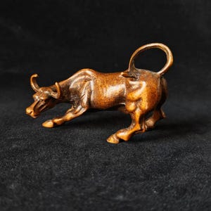 May include: A detailed wooden sculpture of a bull in a charging pose. The bull is brown with visible wood grain and has curved horns and a raised tail. The sculpture is set against a dark background, highlighting the details of the carving.