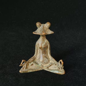 May include: A bronze frog figurine in a seated yoga pose. The frog has a detailed head and body, with its legs crossed and arms resting on its knees. The figurine is set against a dark background.