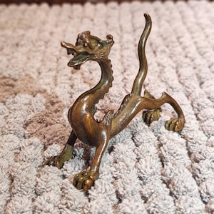 K&ouml;nnte beinhalten: Bronzefarbene Drachenfigur mit offenem Maul und detaillierten Schuppen. Der Drache ist mit ausgestreckten Vorderbeinen und aufw&auml;rts gebogenem Schwanz dargestellt. Auf einer strukturierten, hellgrauen Oberfl&auml;che.