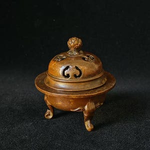 May include: A small, antique bronze incense burner with a lid and three small feet. The lid has decorative cutouts and a small knob on top. The burner is set against a dark background.