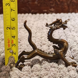 K&ouml;nnte beinhalten: Eine bronzefarbene Drachenfigur, etwa 13 cm lang, mit detaillierten Merkmalen. Der Drache ist in einer dynamischen Pose dargestellt, mit gebogenem K&ouml;rper und erhobenem Kopf. Die Figur steht vor einem strukturierten Hintergrund.