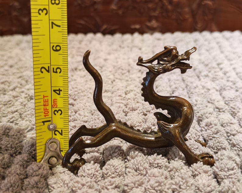 K&ouml;nnte beinhalten: Eine bronzefarbene Drachenfigur, etwa 13 cm lang, mit detaillierten Merkmalen. Der Drache ist in einer dynamischen Pose dargestellt, mit gebogenem K&ouml;rper und erhobenem Kopf. Die Figur steht vor einem strukturierten Hintergrund.
