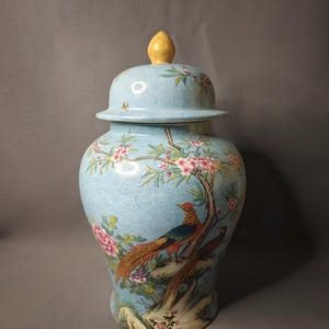 May include: A light blue ceramic jar with a lid, featuring a detailed painting of a pheasant perched on a tree branch with pink blossoms. The jar has a textured surface and a yellow finial on the lid.