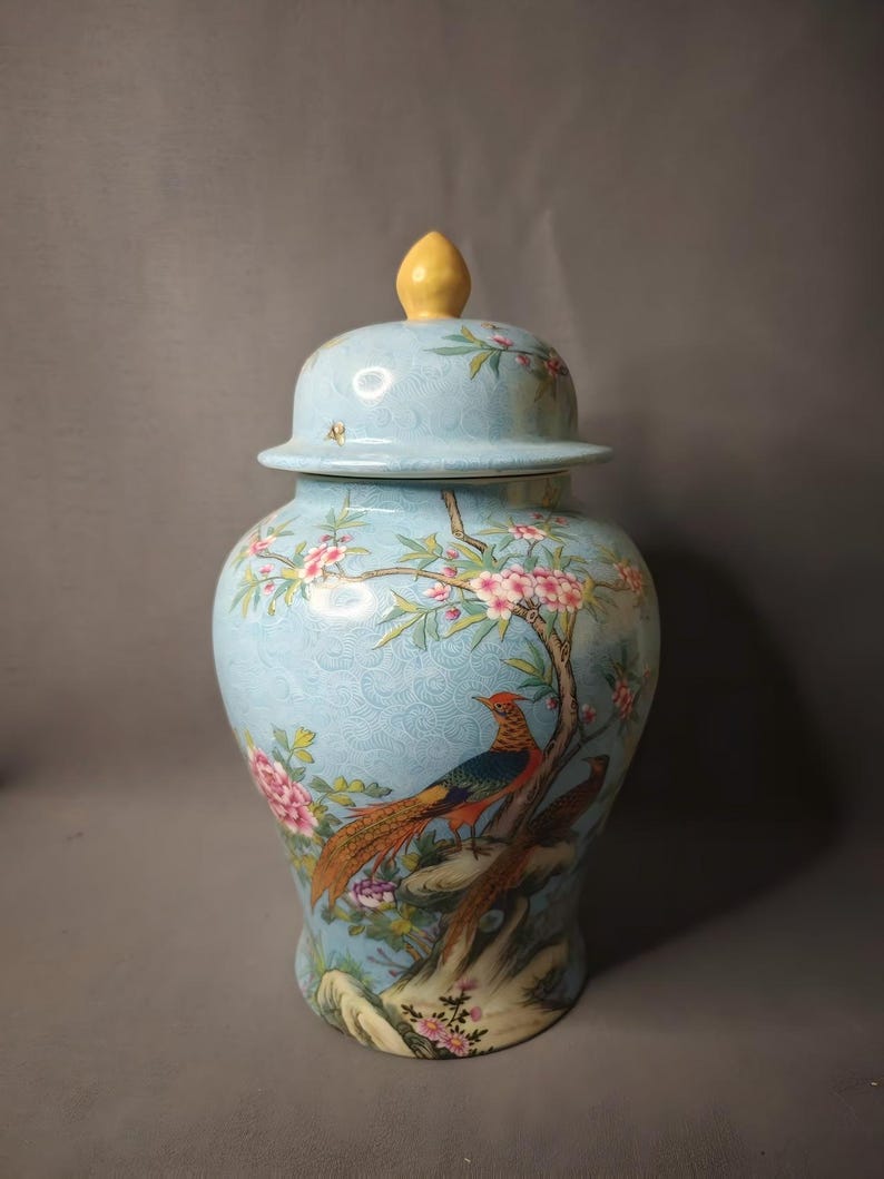 May include: A light blue ceramic jar with a lid, featuring a detailed painting of a pheasant perched on a tree branch with pink blossoms. The jar has a textured surface and a yellow finial on the lid.