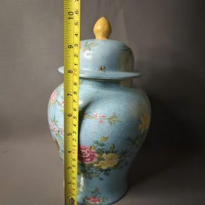May include: A light blue ceramic jar with a lid, adorned with floral patterns in pink, yellow, and green. The jar is approximately 10 inches tall, measured with a yellow tape measure. The lid has a yellow finial.