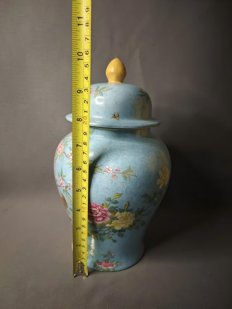 May include: A light blue ceramic jar with a lid, adorned with floral patterns in pink, yellow, and green. The jar is approximately 10 inches tall, measured with a yellow tape measure. The lid has a yellow finial.