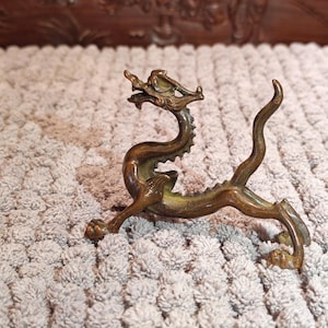 K&ouml;nnte beinhalten: Eine bronzene Drachenfigur mit aufwendigen Details, darunter ein detaillierter Kopf, ein stacheliger R&uuml;cken und ein geschwungener Schwanz. Der Drache ist in einer dynamischen Haltung mit ausgestreckten Beinen dargestellt. Die Figur steht auf einer strukturierten Oberfl&auml;che.