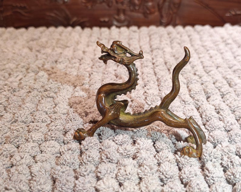 K&ouml;nnte beinhalten: Eine bronzene Drachenfigur mit aufwendigen Details, darunter ein detaillierter Kopf, ein stacheliger R&uuml;cken und ein geschwungener Schwanz. Der Drache ist in einer dynamischen Haltung mit ausgestreckten Beinen dargestellt. Die Figur steht auf einer strukturierten Oberfl&auml;che.