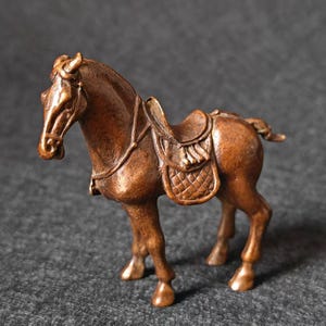 Könnte beinhalten: Bronzefarbene Pferdfigur mit Sattel. Das Pferd steht auf vier Beinen, den Kopf erhoben. Der Sattel ist mit einem Steppmuster versehen. Die Figur ist wahrscheinlich aus Metall und ein dekoratives Objekt.