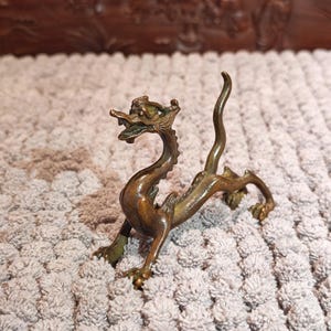 K&ouml;nnte beinhalten: Eine bronzefarbene Drachenfigur mit detailliertem Design. Der Drache ist mit erhobenem Kopf und gebogenem K&ouml;rper dargestellt. Die Figur besteht aus einem metallischen Material und hat ein verwittertes Aussehen. Der Drache ist ein dekoratives Objekt.